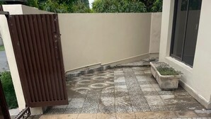 Un espace extérieur pavé, avec une entrée fermée par un portail, un bac à fleurs en béton et un mur percé d’une fenêtre.