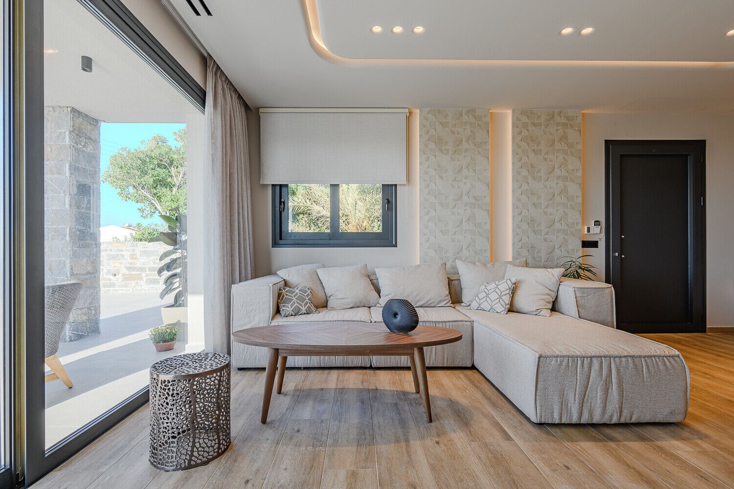 Un salon moderne avec un canapé d’angle, une table basse ronde en bois et une grande fenêtre donnant sur l’extérieur.