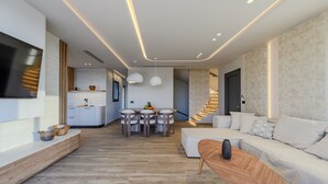A modern living room with a sofa, a wooden coffee table, and a TV mounted on the wall. There is a dining area with a table and chairs, and a kitchen in the background. - Nysa Luxury 2 (Hersonissos)