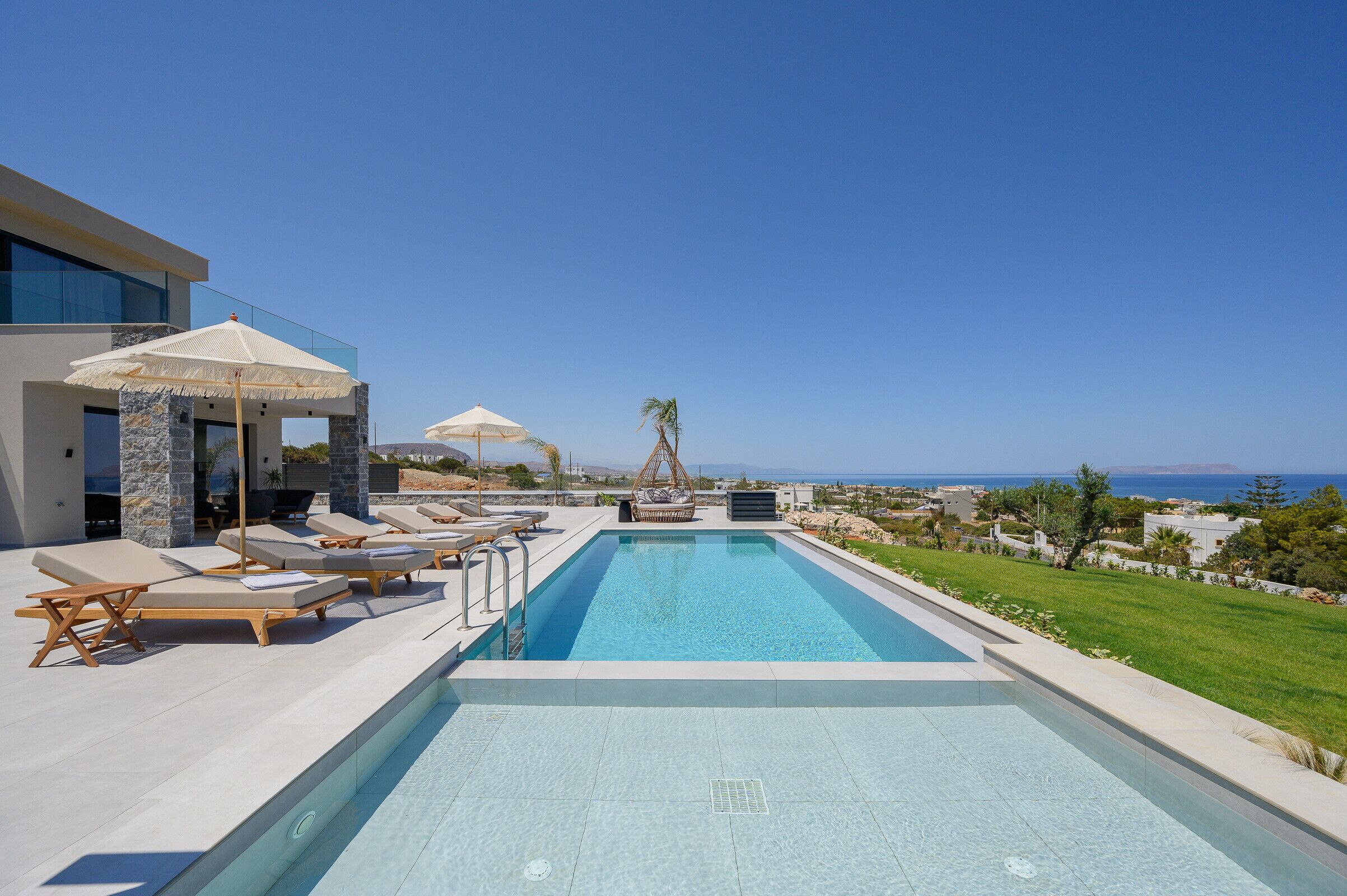 A pool area with lounge chairs and umbrellas, a clear view of the sea, and a modern building with stone and glass elements.