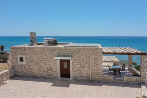A stone building with a door and a small window, a pergola with a table and chairs, and a view of the sea.