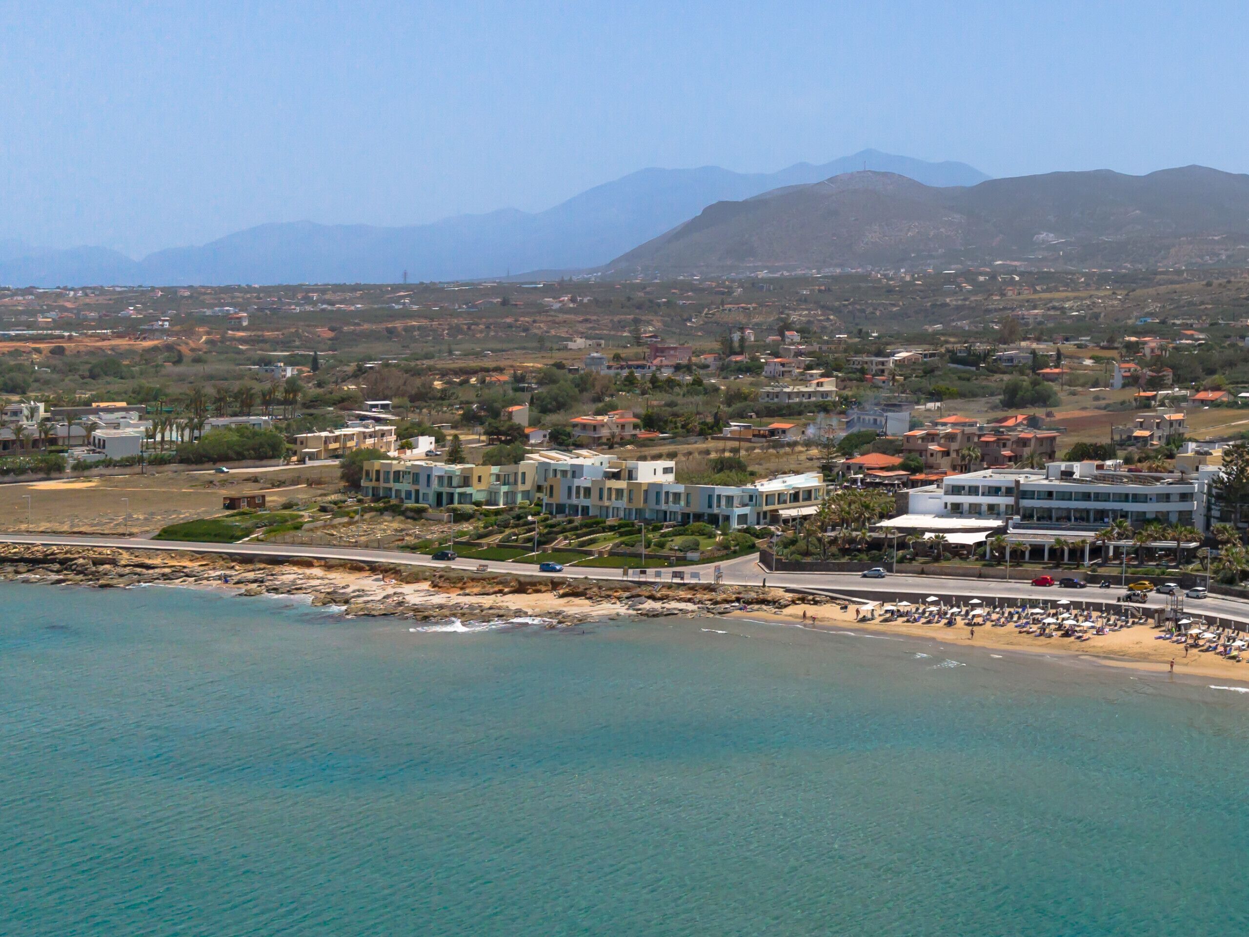 A coastal town with modern buildings, a beach, and mountains in the background.