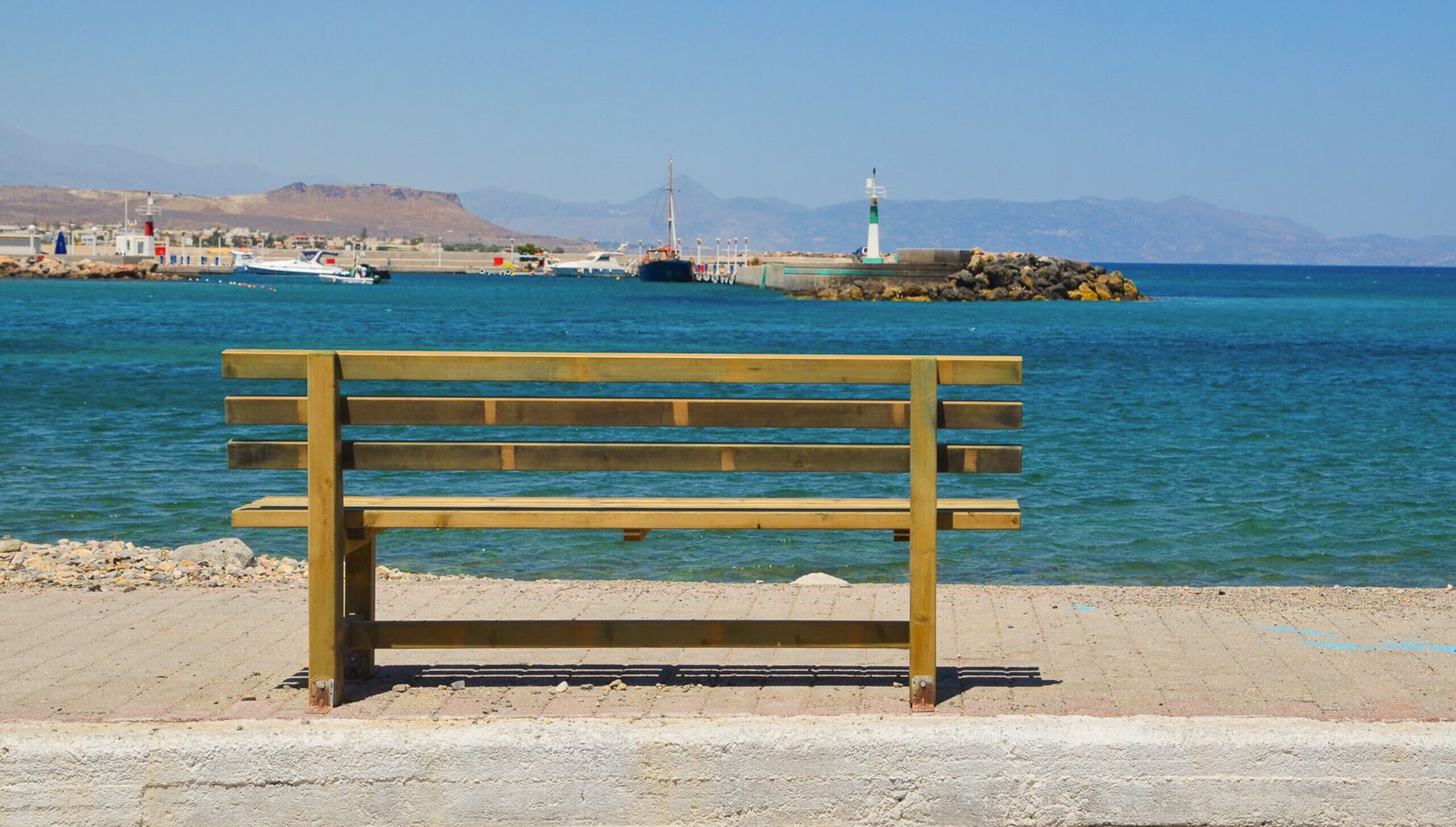 A wooden bench by a waterfront with boats and a lighthouse in the background.