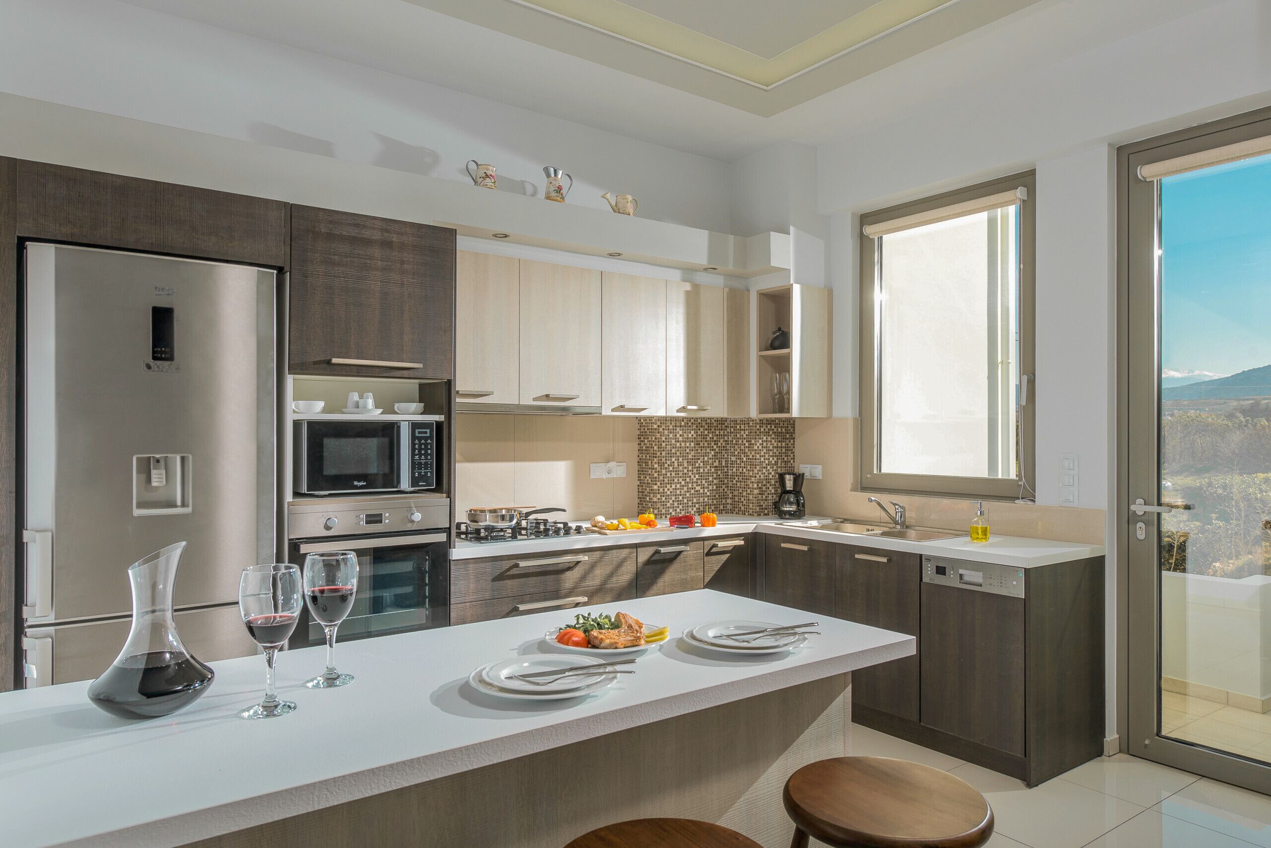 Une cuisine moderne dotée d’un grand îlot central, d’appareils électroménagers en acier inoxydable et offrant une vue sur l’extérieur.