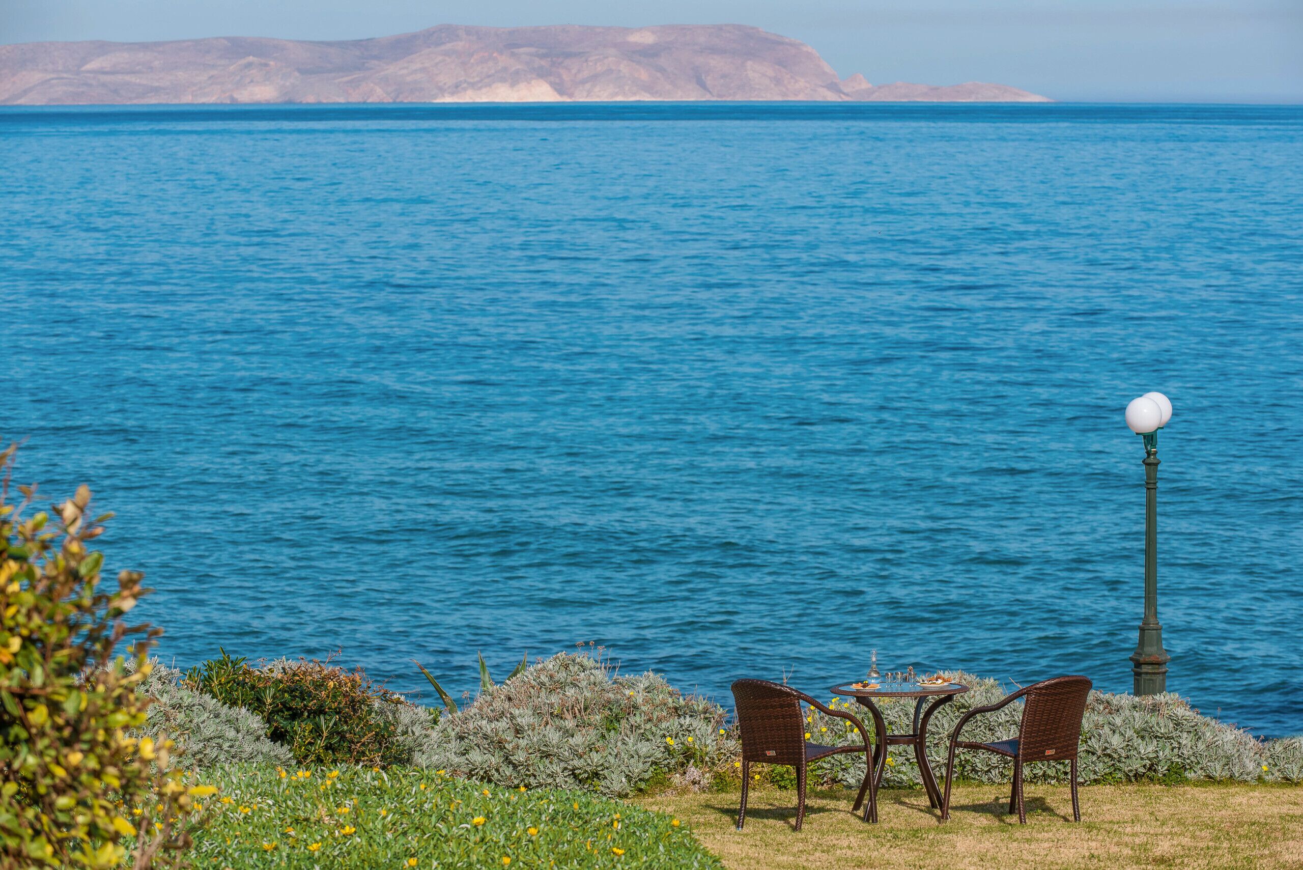 Une vue sur la mer avec une table et des chaises au bord de l’eau, un lampadaire et une chaîne de montagnes à l’horizon.