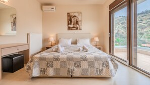 A bedroom with a bed, two bedside tables, a nightstand, a dresser, a mirror, and a large window with a view of a hilly landscape. - Villa Nikos (Malevizi)
