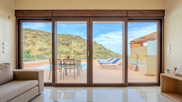 A modern living room with a large sliding glass door leading to a pool area with lounge chairs and a table. - Villa Nikos (Malevizi)