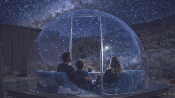 Une structure en forme de dôme transparent offrant une vue dégagée sur le ciel nocturne, une terrasse en bois et un espace détente avec des coussins.