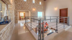 A spacious interior with a stone wall, a balcony with a railing, and a dining area with tables and chairs. - Arios Lux Villa (Rethymno)