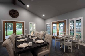 A dining area with a long table set for six, surrounded by white chairs, with large windows and doors providing views of the outdoors. - Vineyard Cottage #2 with Complimentary Breakfast (Gillespie County)