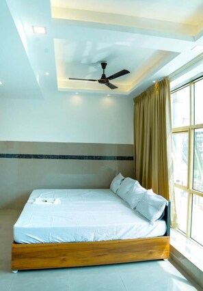 A neatly made bed with white linens in a room with a ceiling fan, large windows with curtains, and a tiled floor. - Nearest Rooms (Thrissur)