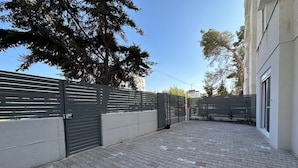A paved courtyard with a metal gate, a building with a window, and trees in the background.