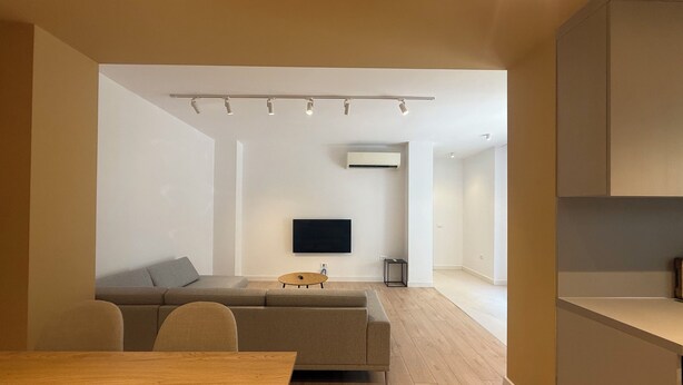 A modern living room with a sofa, a wooden dining table, and a flat-screen TV mounted on the wall.