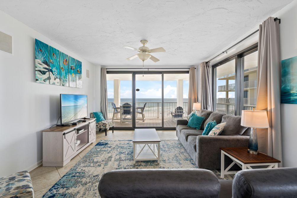 A spacious living room with a large TV, comfortable seating, a ceiling fan, and a view of the ocean through the sliding glass doors.
