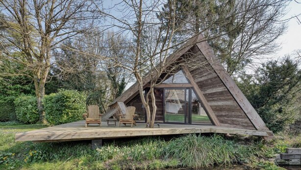 A wooden cabin with a sloped roof, a deck with two chairs, and a glass door leading inside.