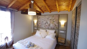 A bedroom with a wooden ceiling, a bed with white bedding, a wall-mounted light, and a floral painting.