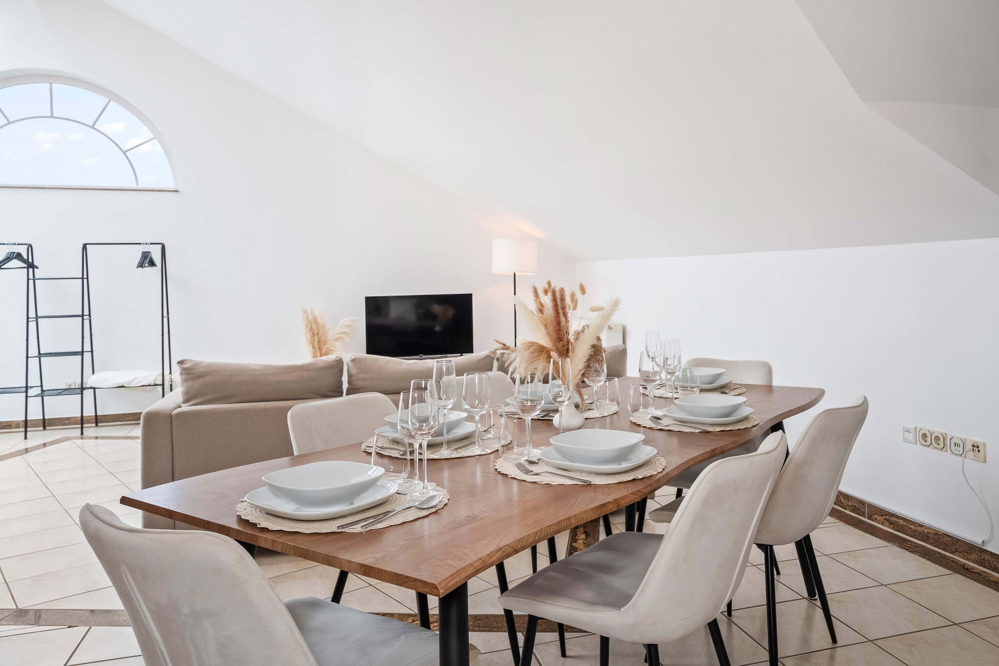 Ein Essbereich mit einem Holztisch, gedeckt für sechs Personen, umgeben von weißen Stühlen, einer Couch und einem Fernseher.