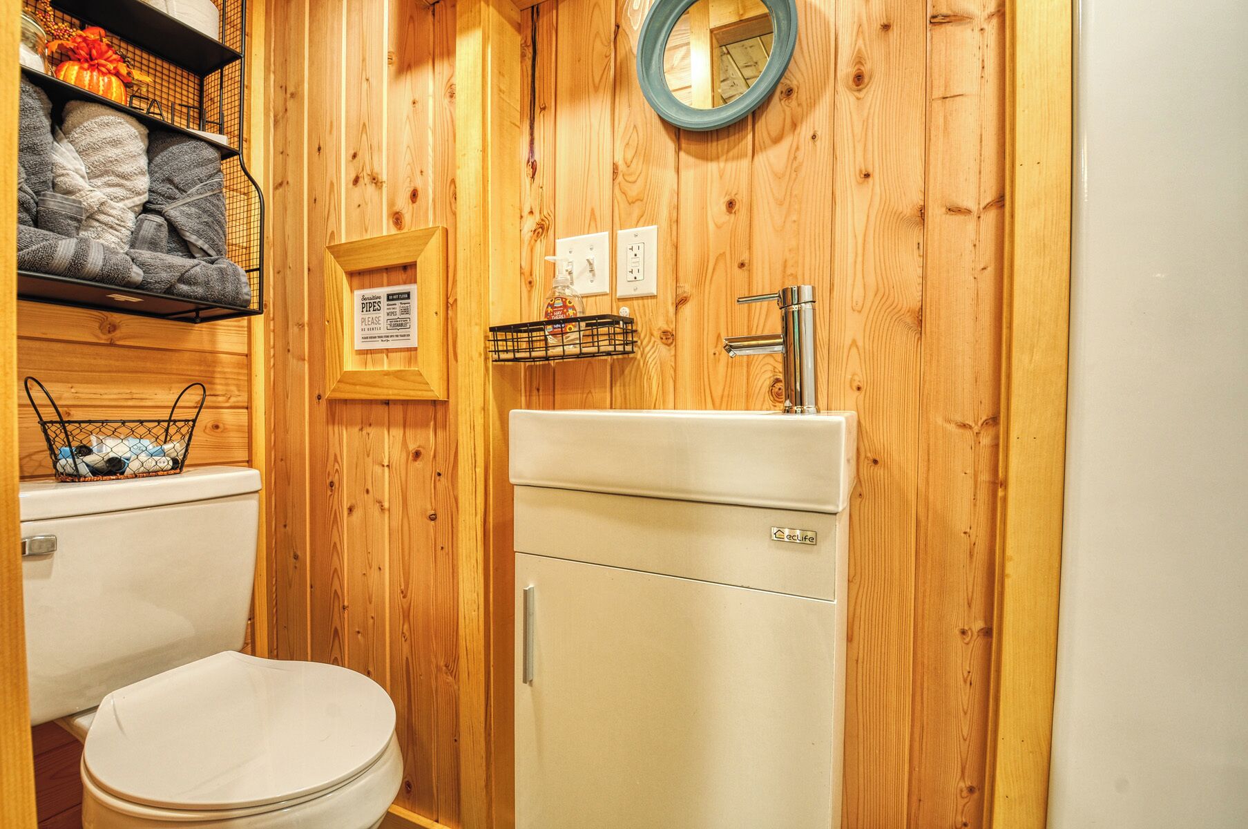 A compact bathroom with a wooden wall, a toilet, a sink with a mirror, and a shelf with towels and toiletries.