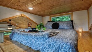 A bedroom with a wooden ceiling, a bed with a blue floral bedspread, a window with a view of greenery, and a small table with fruits and a decorative item.