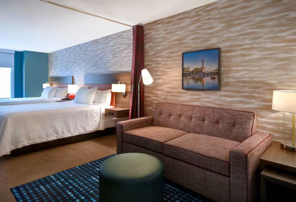 A hotel room with a bed, a sofa, a nightstand, a lamp, and a painting on the wall.