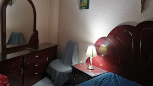 A bedroom with a bed, a desk with a lamp, a chair, a dresser, a mirror, and a wall decoration.