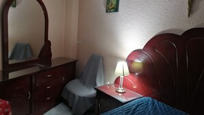 Un dormitorio con cama, una silla, escritorio con lámpara, cómoda, espejo y decoración en la pared.