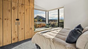 A bedroom with a large wooden wardrobe, a bed with a beige blanket, and a view of a house and mountains through a large window. - Eight On Jackson - Luggate Holiday Home (Cromwell)