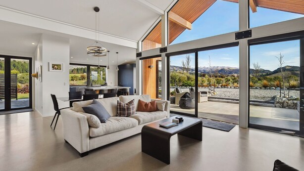 A spacious living room with a large sofa, a coffee table, and a view of a mountainous landscape through large windows. - Eight On Jackson - Luggate Holiday Home (Cromwell)