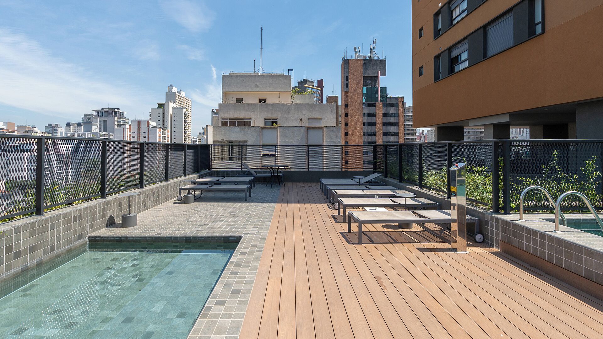A rooftop pool area with lounge chairs and a small pool, surrounded by a fence and city buildings in the background.