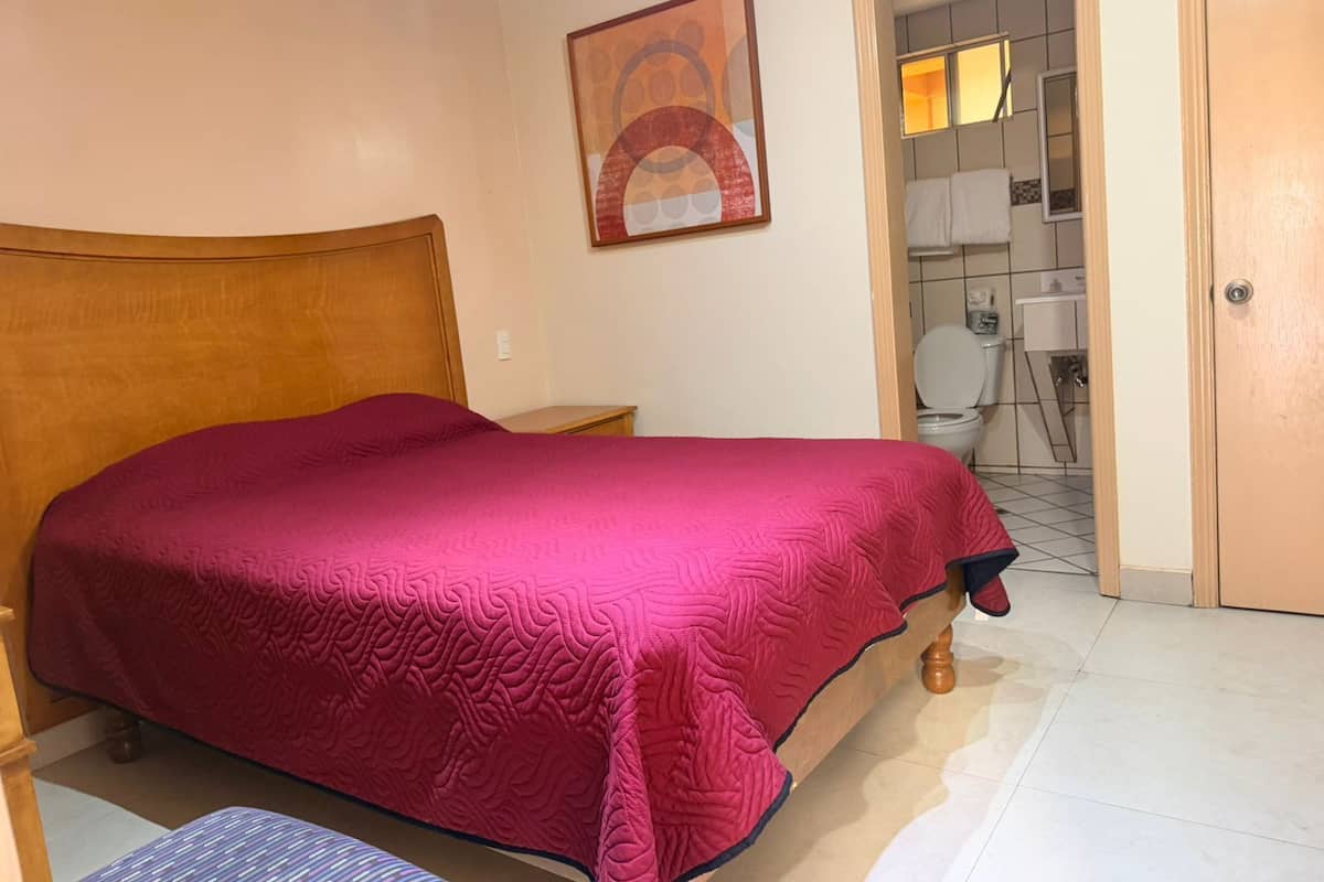 Un dormitorio con cama, mesita de noche, silla, baño visible a través de una puerta abierta y un cuadro en la pared.