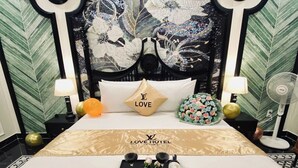A hotel room with a bed, a headboard with a floral design, a bedside table with a lamp, a decorative pillow, and a tray with wine glasses and a bottle.