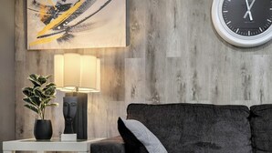 A modern living room with a grey sofa, a white side table, a black lamp, a potted plant, a minimalist sculpture, and a large abstract painting. - Modern 2BR Condo with secure parking and walking distance to Rogers Place  (Edmonton)