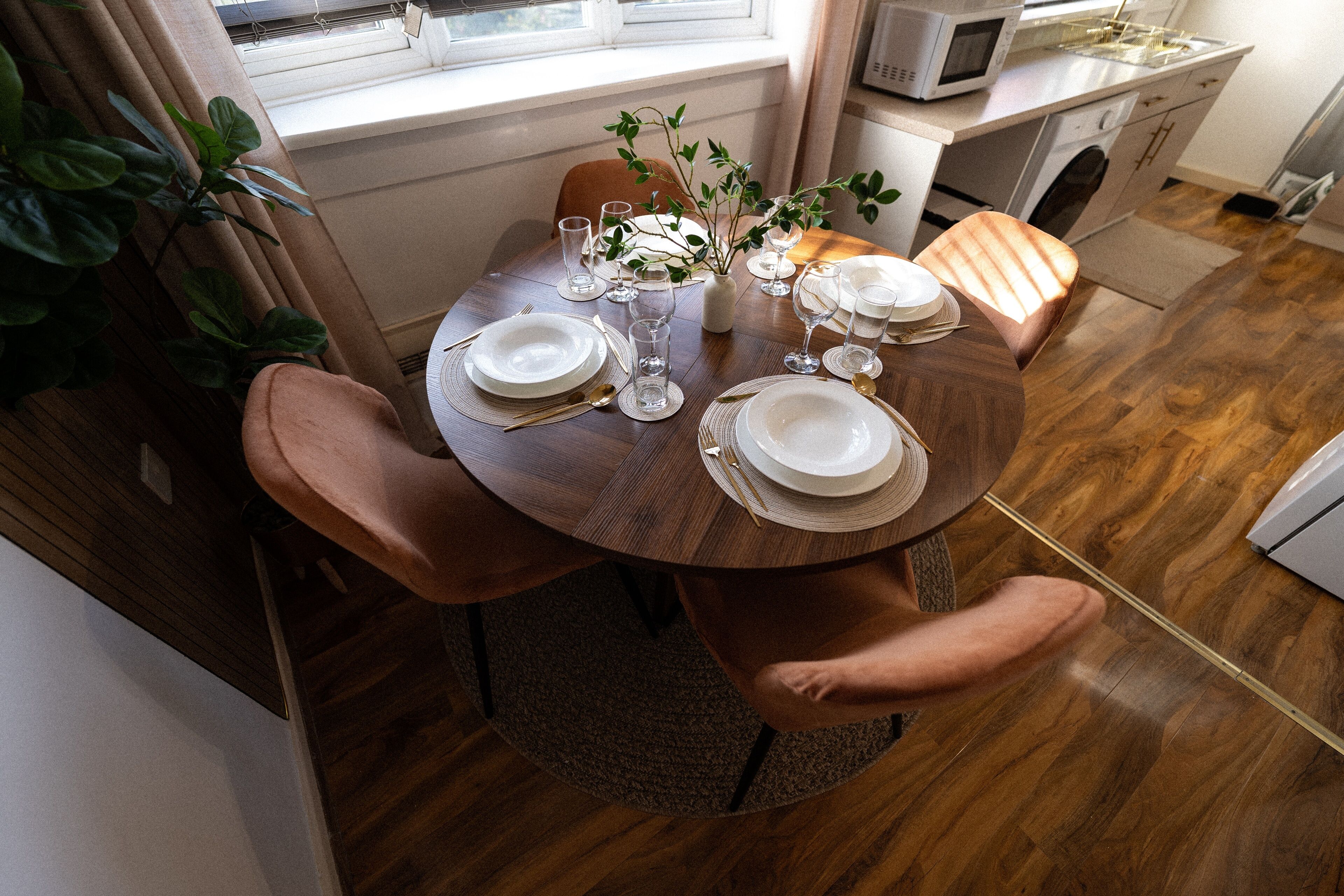 A dining table set for four with white plates, glasses, and cutlery. There are two chairs, a vase with greenery, and a window above the table.