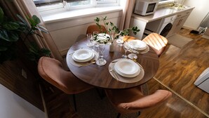 A dining table set for four with white plates, glasses, and cutlery. There are two chairs, a vase with greenery, and a window above the table.