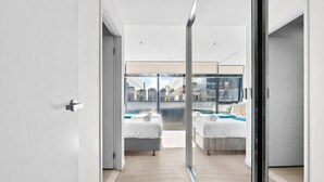 A modern hotel room with a glass door leading to a balcony, two beds, and a city view.