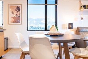 Zona pranzo moderna con tavolo rotondo, sedie bianche, vista sulla città tramite ampie finestre e un quadro appeso al muro.