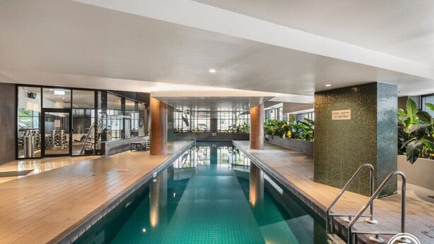 Una piscina con suelo de baldosas, rodeada por una terraza de madera y paredes de vidrio.