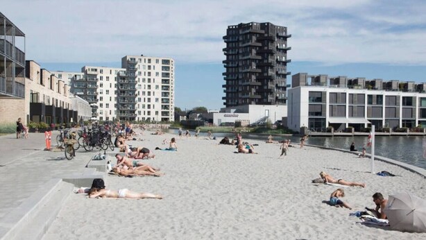Ein Strand mit badenden und sonnenbächelnden Menschen, umgeben von modernen Gebäuden und einem Wasserabschnitt.