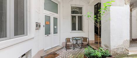 A small courtyard with a tiled floor, two wicker chairs, a round table, a wooden planter with green plants, and a door with a sign.