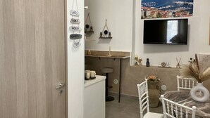 A modern kitchen with a wooden door, a dining area with white chairs, a TV mounted on the wall, and a scenic view of a coastal town.