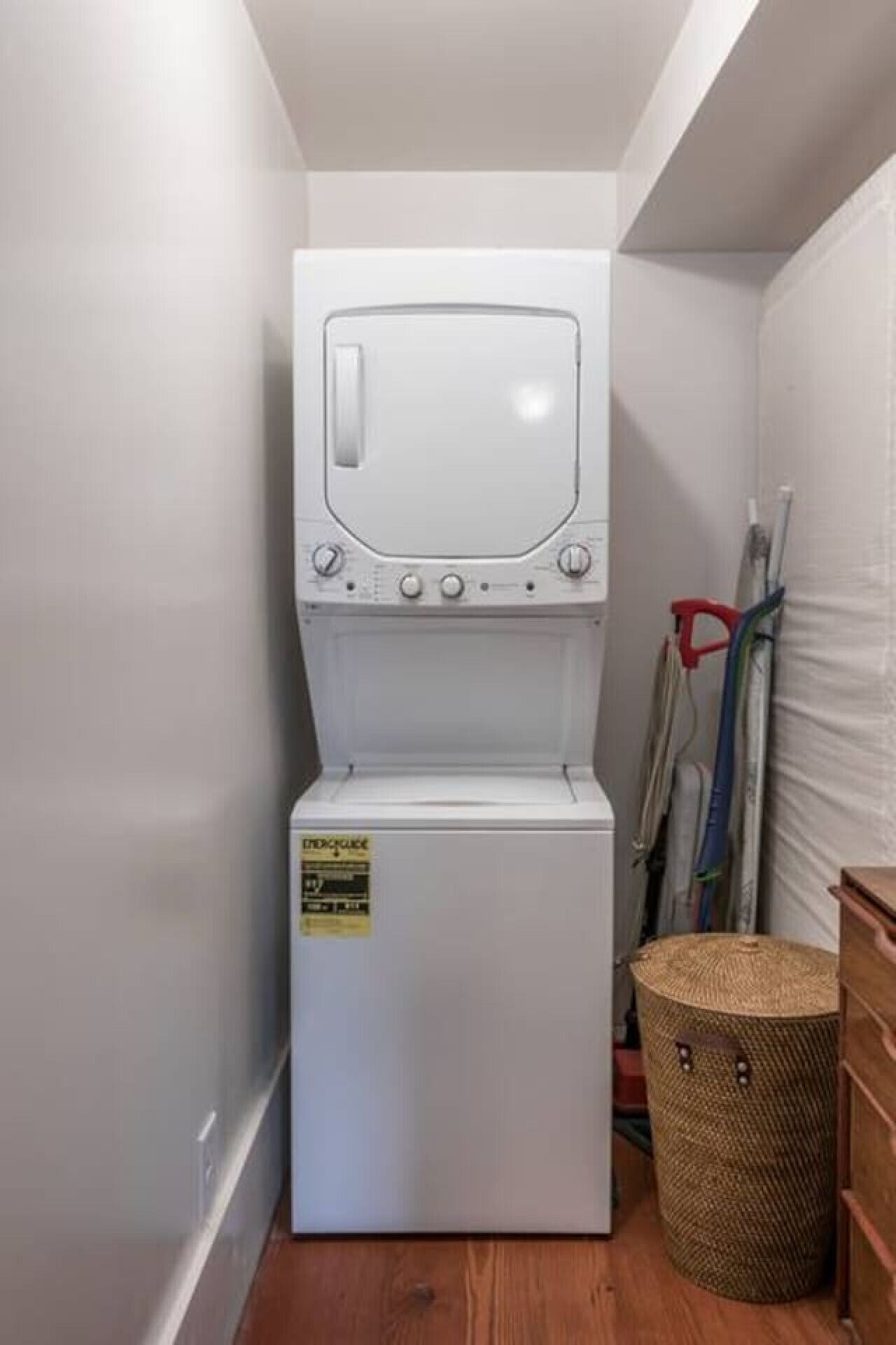 A compact, white laundry machine in a narrow space with a wooden floor, a wicker basket, and a red-handled mop leaning against the wall.