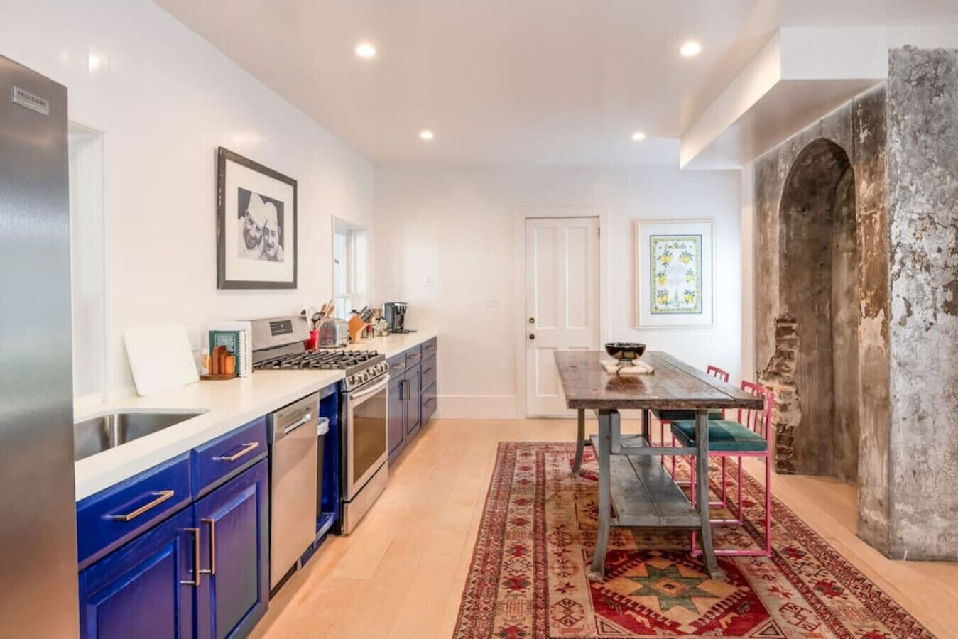 A modern kitchen with a dining area, featuring a large window, a rustic stone wall, and a traditional rug.