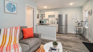 A modern apartment with a grey sofa, a striped throw blanket, a small round coffee table with a plant, a kitchen with white cabinets, a stainless steel refrigerator, and a bar stool.