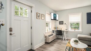 A modern living room with a white door, a small desk with a chair, a mounted TV, and a couch.