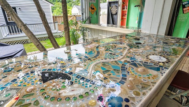 A mosaic-topped table in an outdoor setting with a dartboard and a shelf with items. - Casa de Olas~3br/2ba Home with Backyard Oasis! (North Charleston)
