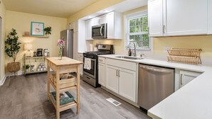 A modern kitchen with white cabinetry, stainless steel appliances, and a wooden table with a vase. - Casa de Olas~3br/2ba Home with Backyard Oasis! (North Charleston)
