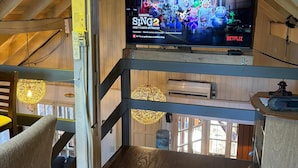 Interior de madera con un televisor mostrando 'Sing 2' en Netflix, un dispensador de agua y techo de madera.