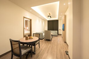 A hotel room with a round dining table, two chairs, a sofa, a television, and a ceiling light fixture.