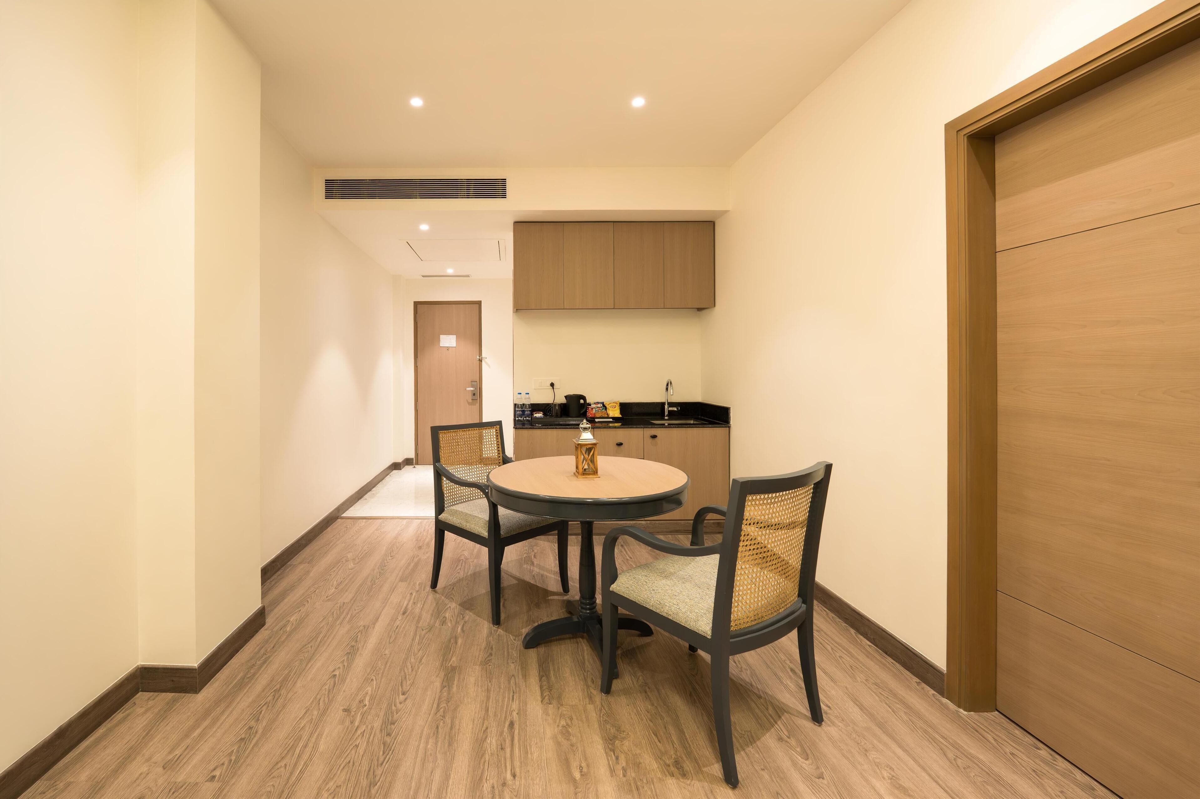 Habitación de hotel con comedor que incluye una mesa redonda y cuatro sillas, zona de cocina con armarios y encimera, y una puerta de madera grande.