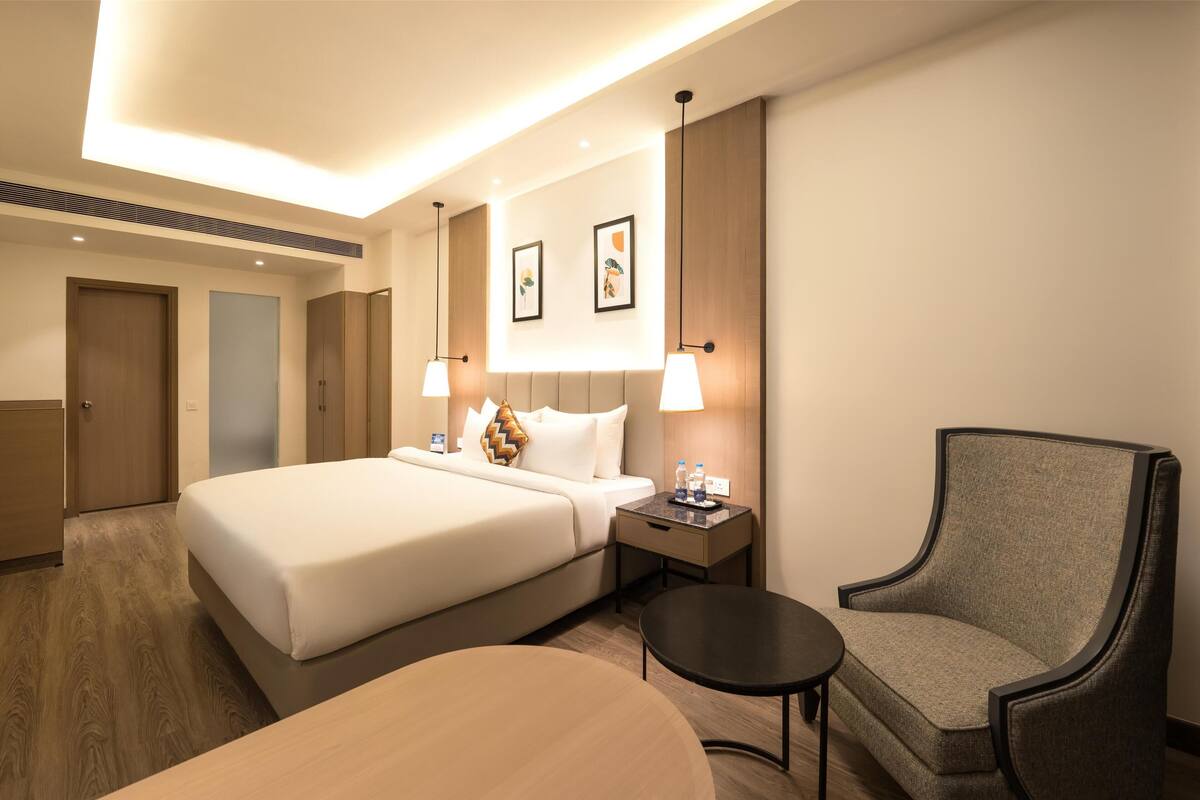 A hotel room with a large bed, a chair, a small table, and a nightstand with a lamp.